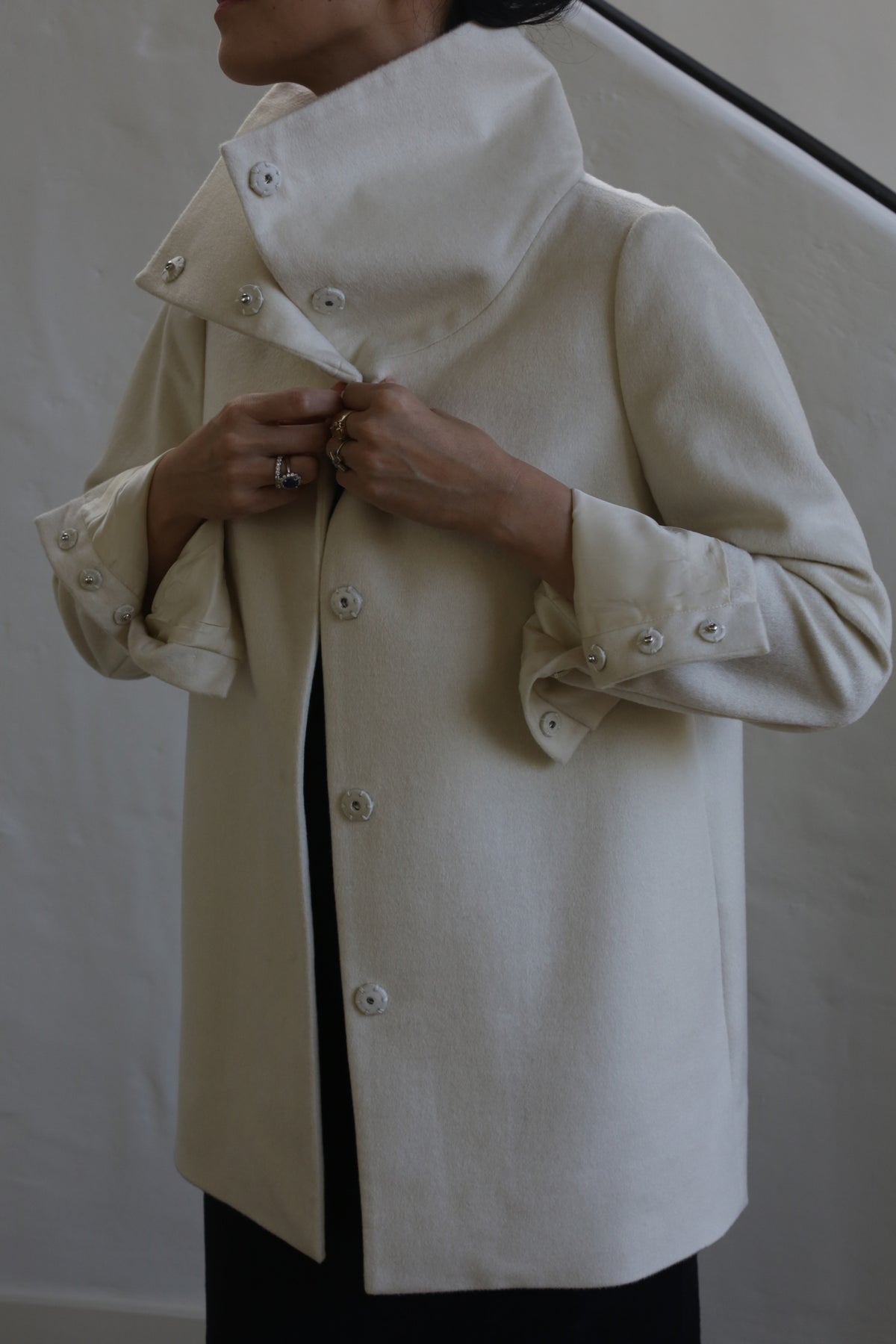 A person wearing the Stamford II | Undyed, a high-collared coat crafted from woven cashmere, adjusts the button-secured cuffs. The scene is set against a simple, light-colored wall and staircase, with the individual's face not visible.