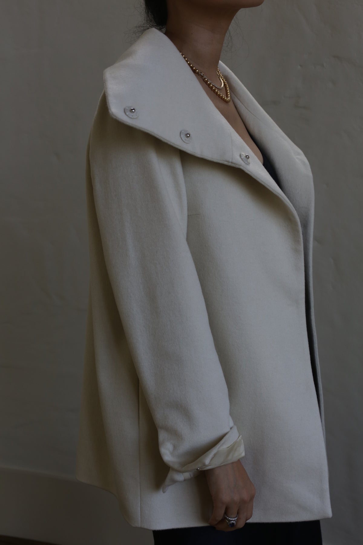 A person is wearing the Stamford II | Undyed, a light-colored, loose-fitting, long-sleeved coat with a high collar. Their face is out of frame, and they are accessorized with a gold necklace and rings on their fingers. The background is plain and muted.