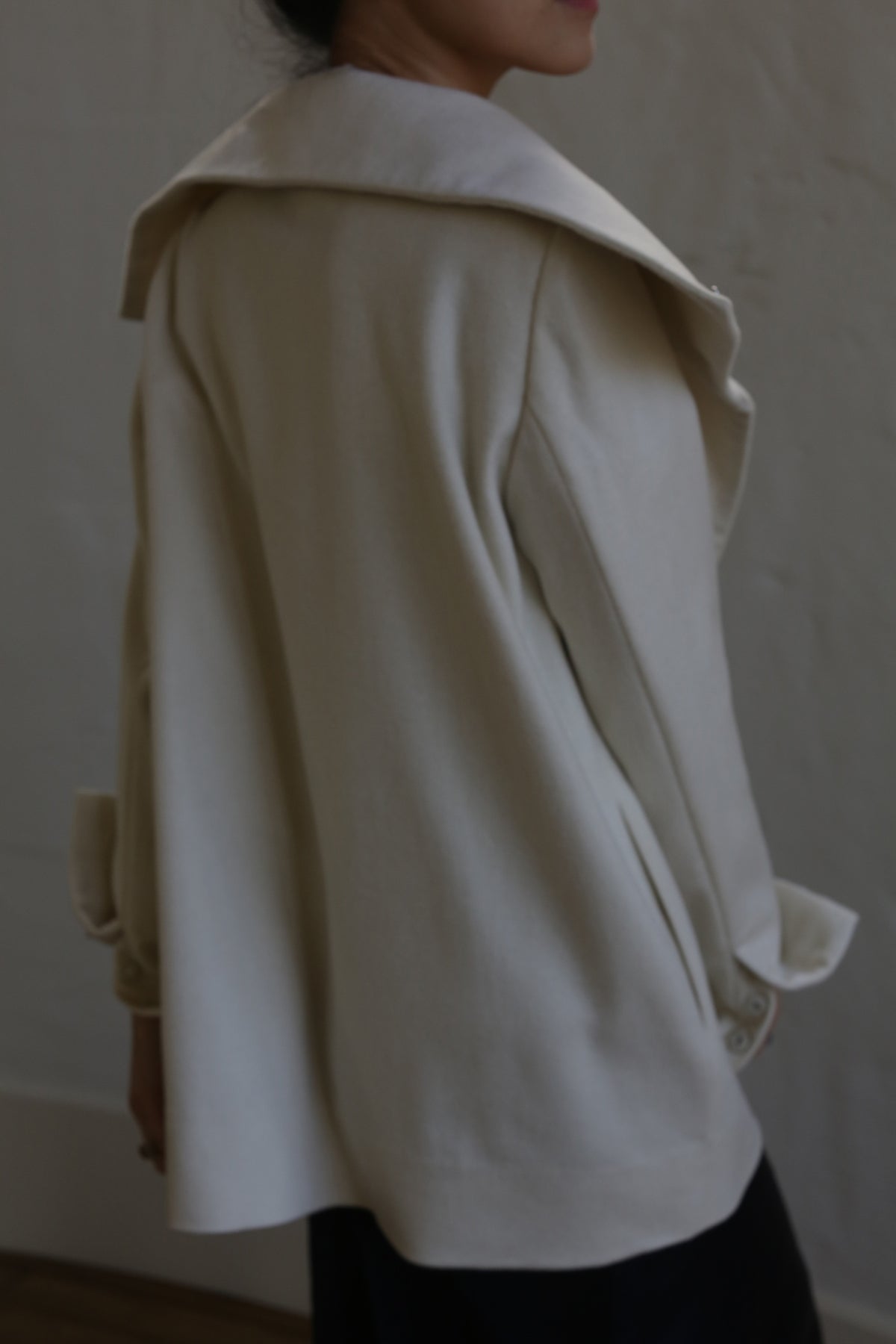 A person stands with their back to the camera, wearing a light-colored, woven Stamford II Undyed cashmere coat with a high collar. The sleeves of the Stamford II coat feature buttoned cuffs. The background is blurred and neutral-colored, emphasizing the individual and the coat.