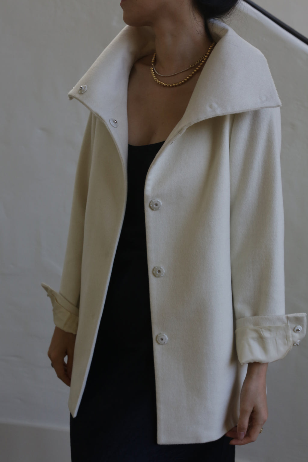 A person is wearing the Stamford II model, a woven cashmere coat in its undyed cream color, featuring large buttons and turned-up cuffs over a black garment. The high-collar coat is complemented by a gold necklace. The individual's head is turned to the side against a plain, light-colored wall.
