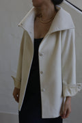 A person is wearing the Stamford II model, a woven cashmere coat in its undyed cream color, featuring large buttons and turned-up cuffs over a black garment. The high-collar coat is complemented by a gold necklace. The individual's head is turned to the side against a plain, light-colored wall.