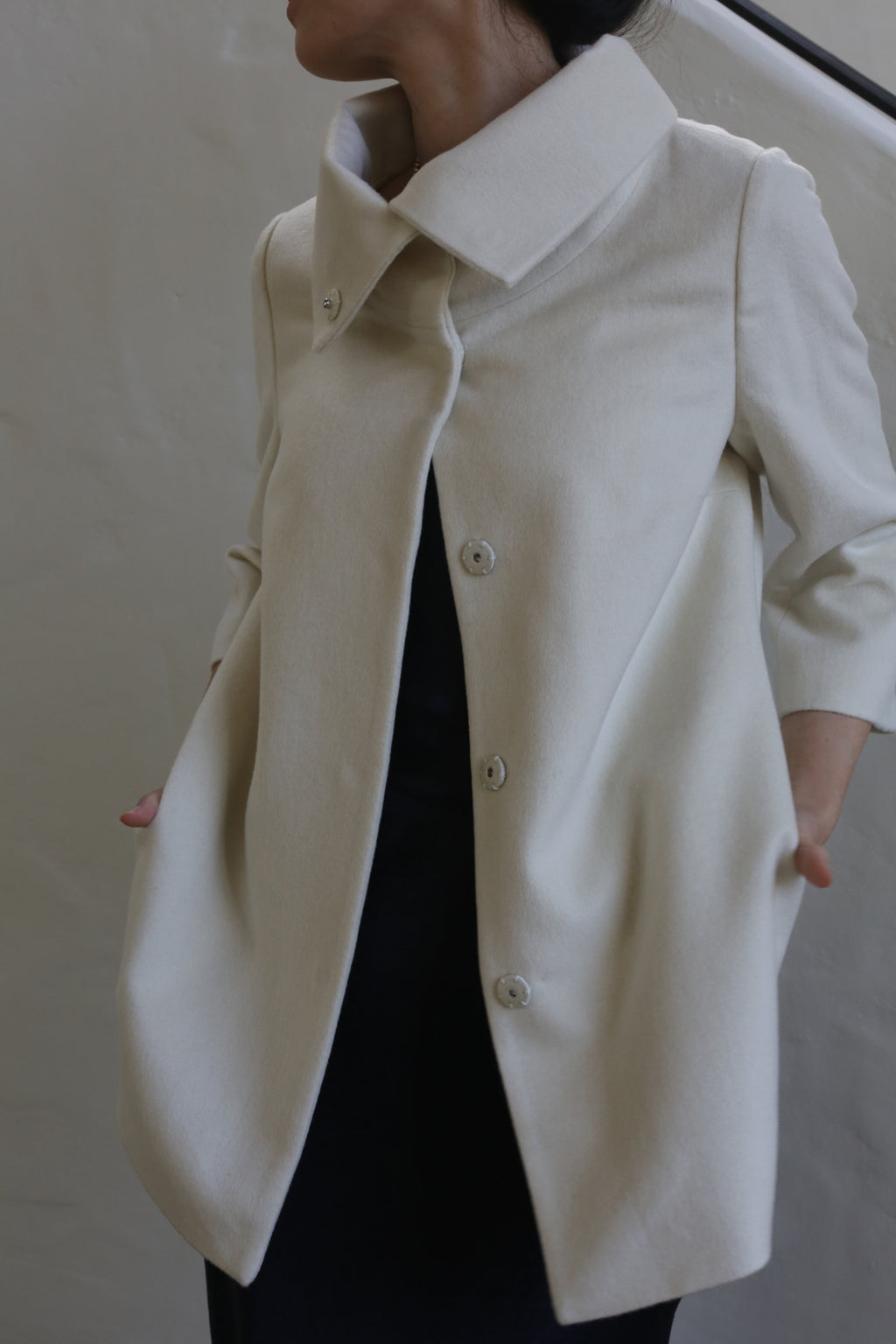A person wears the Stamford | Undyed, a cream oversized coat with large buttons and a high collar, made from undyed woven cashmere, standing against a plain light background with their face partially out of view.