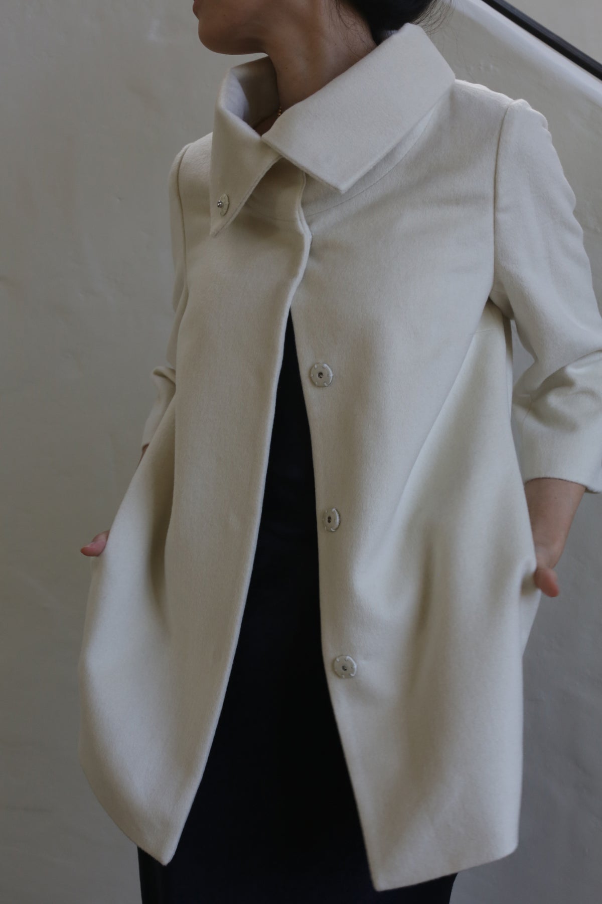 A person wears the Stamford | Undyed, a cream oversized coat with large buttons and a high collar, made from undyed woven cashmere, standing against a plain light background with their face partially out of view.