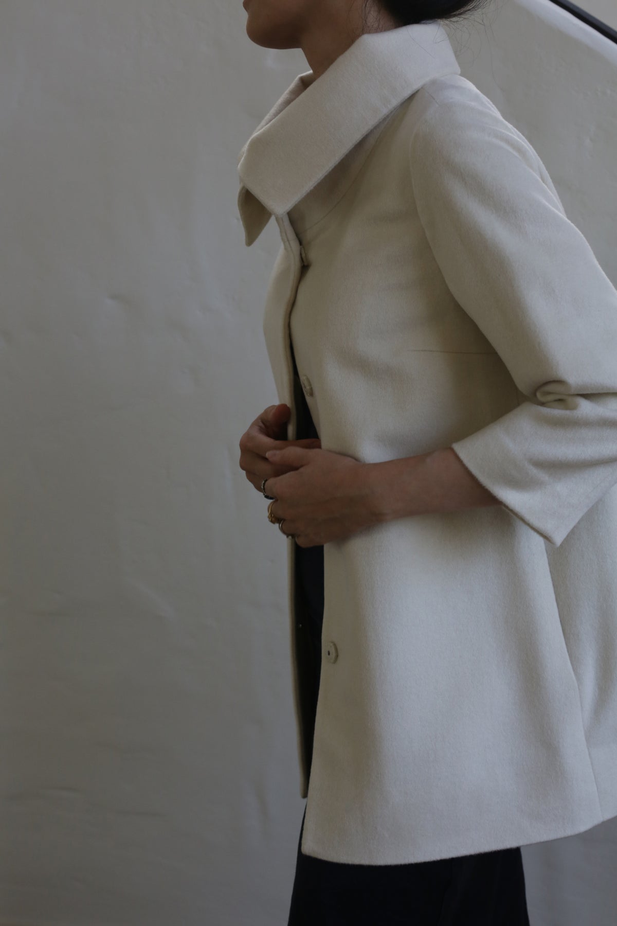 Wearing the Stamford | Undyed, a high-collar, long-sleeve coat in undyed woven cashmere, a person stands against a plain background with hands clasped at the front. Their head is partially out of frame, highlighting the coat’s understated elegance.