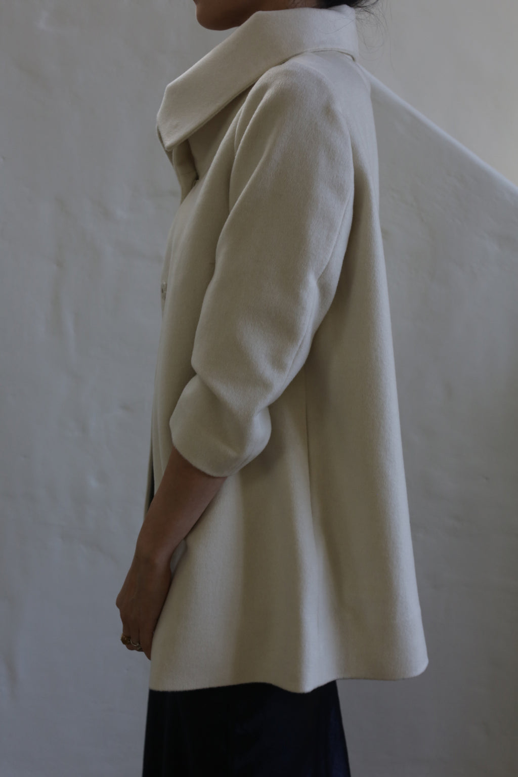 A person stands sideways wearing the Stamford | Undyed woven cashmere coat with a high collar, arms relaxed by their side, against a plain textured wall.
