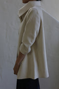 A person stands sideways wearing the Stamford | Undyed woven cashmere coat with a high collar, arms relaxed by their side, against a plain textured wall.