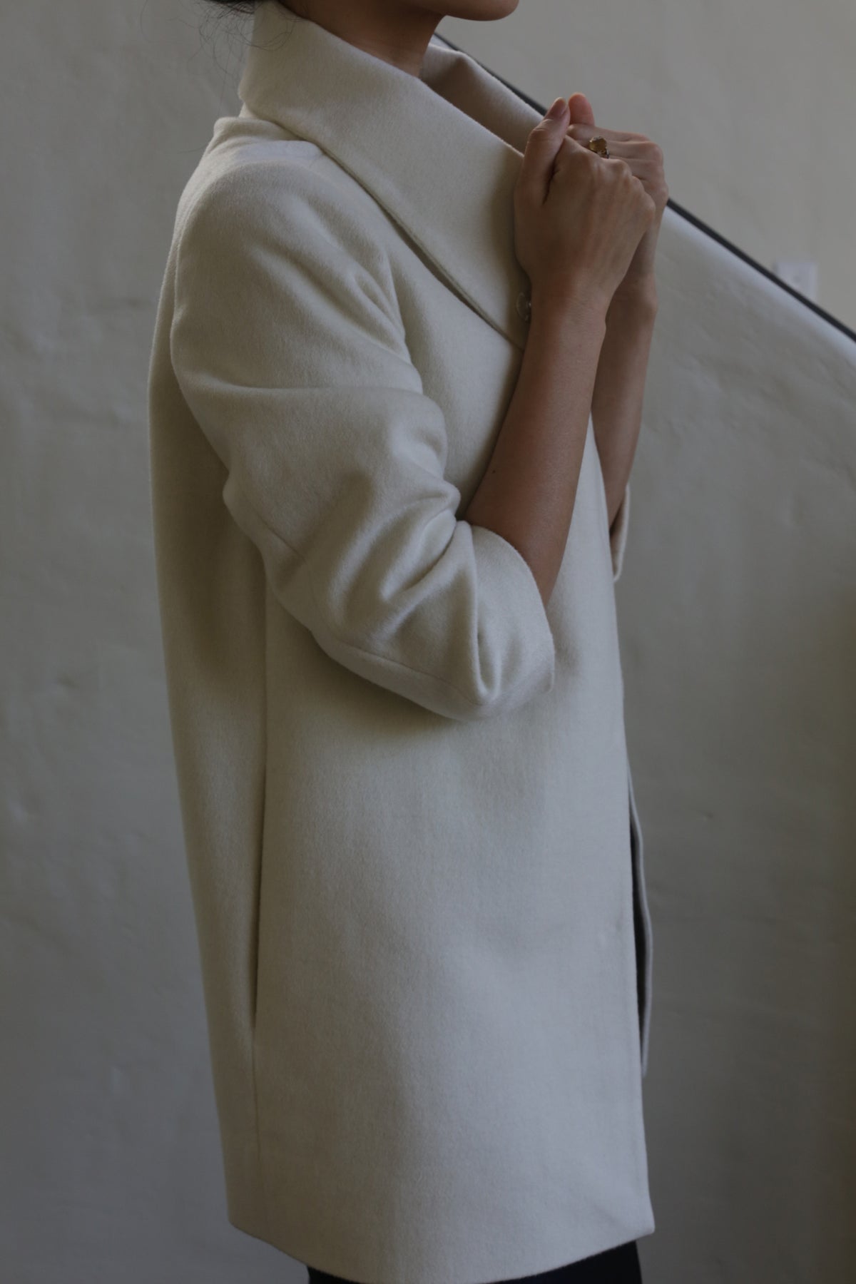 Someone stands against a plain background, gently holding the high collar of the Stamford | Undyed woven cashmere coat, their face partially out of view.