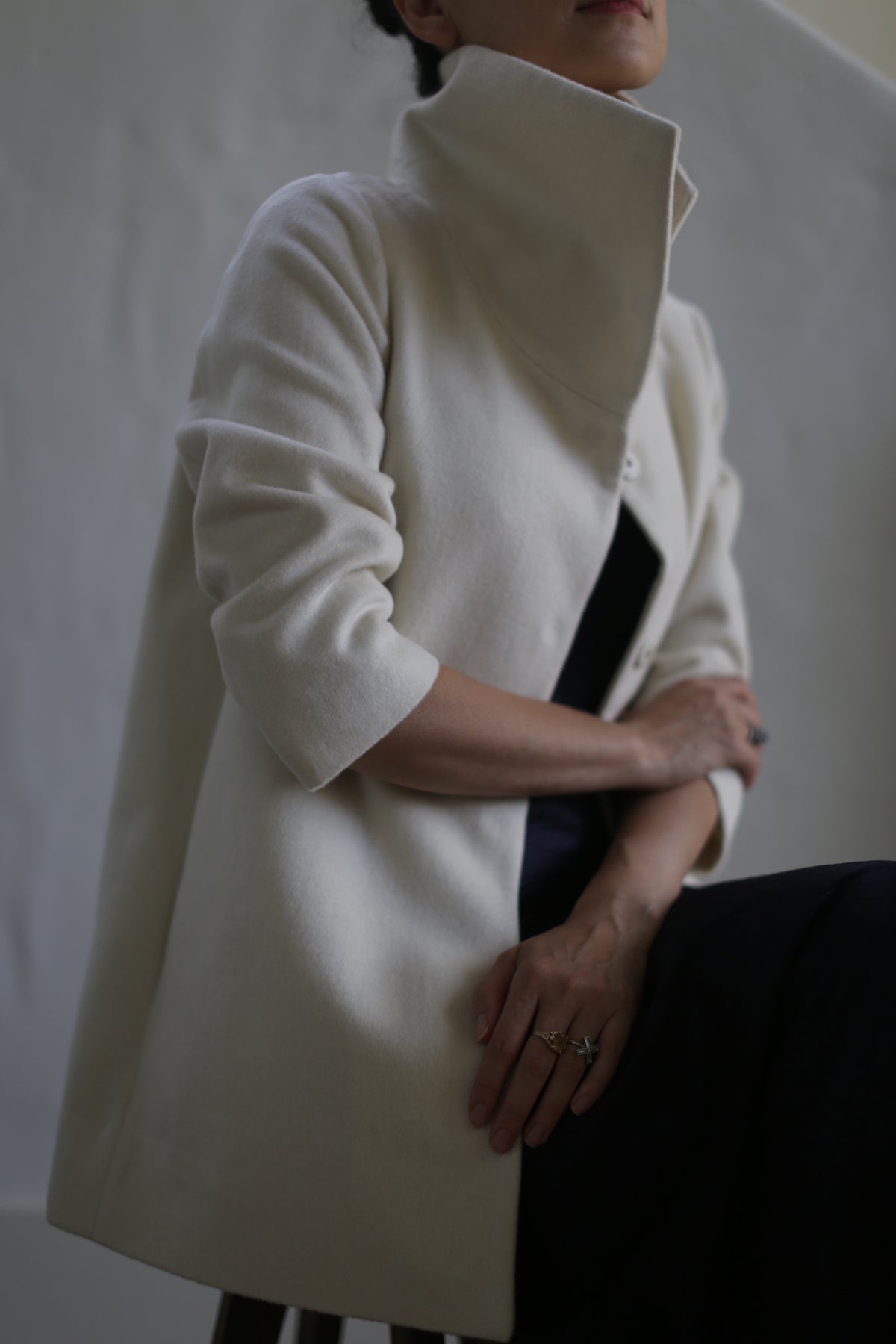 Wearing the Stamford | Undyed woven cashmere coat with a high collar and 3/4 sleeves pushed up, a person sits with arms crossed and hands on their knee against a neutral background, their face partially out of frame.