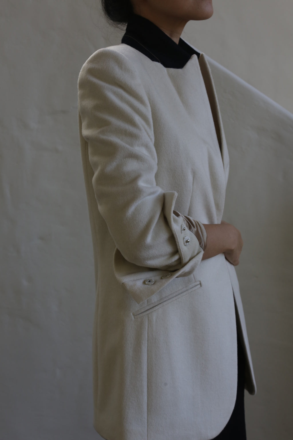 A person stands against a neutral background, wearing the Sloane | Cashmere Undyed blazer that embodies the precision of men's tailoring. It features rolled-up sleeves and a black collar. Their arms are crossed and head is turned to the side, not fully visible.