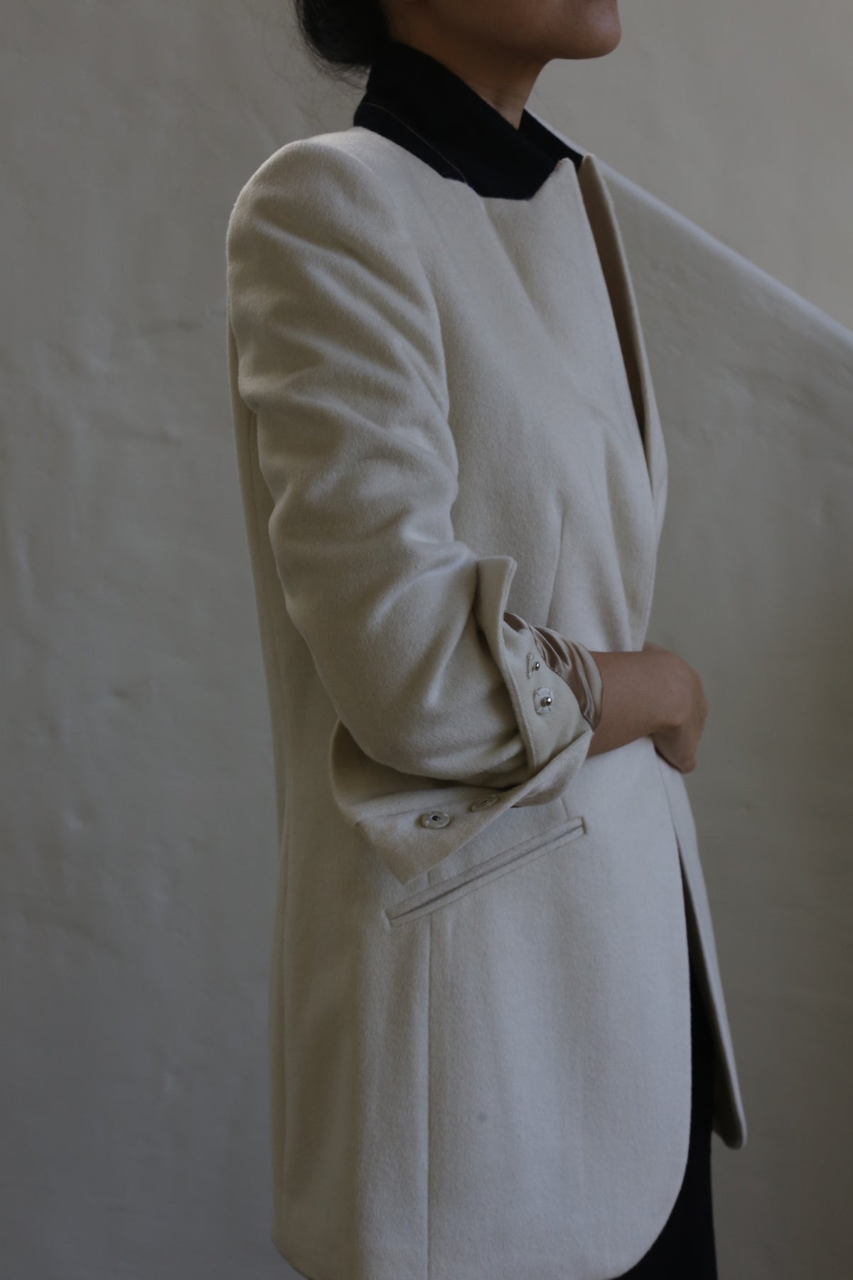 A person stands against a neutral background, wearing the Sloane | Cashmere Undyed blazer that embodies the precision of men's tailoring. It features rolled-up sleeves and a black collar. Their arms are crossed and head is turned to the side, not fully visible.