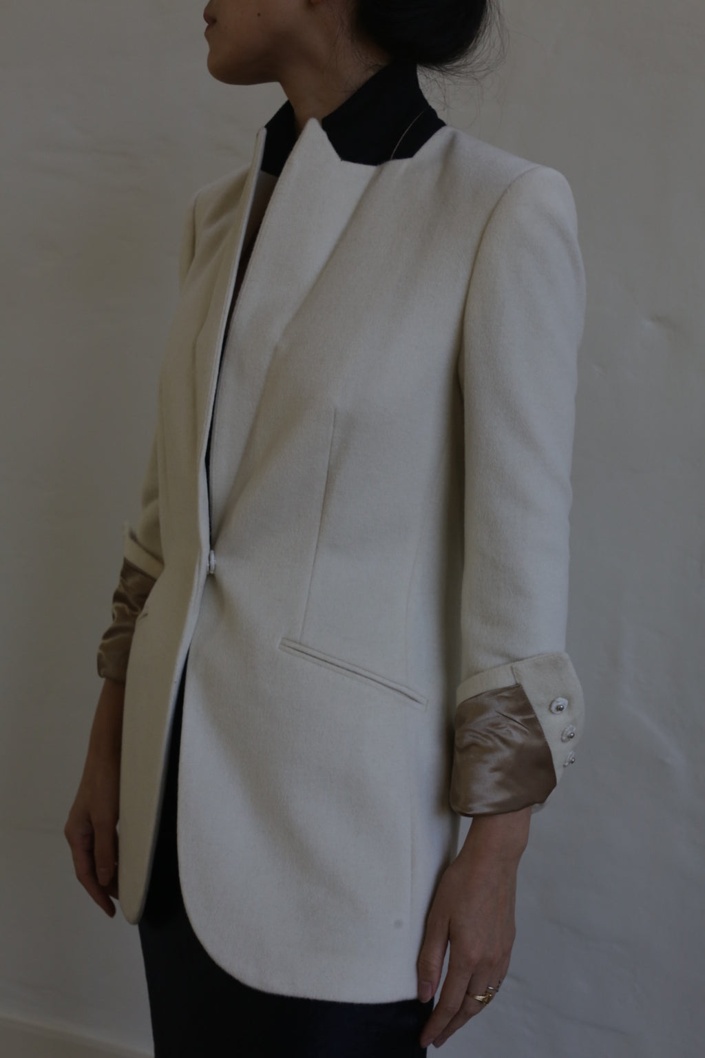 A person wearing the Sloane cashmere undyed blazer, tailored in a cream color with metallic sleeve cuffs, stands in profile against a plain backdrop. The blazer features a sleek design reminiscent of men's tailoring, complete with a concealed front closure and a side pocket.