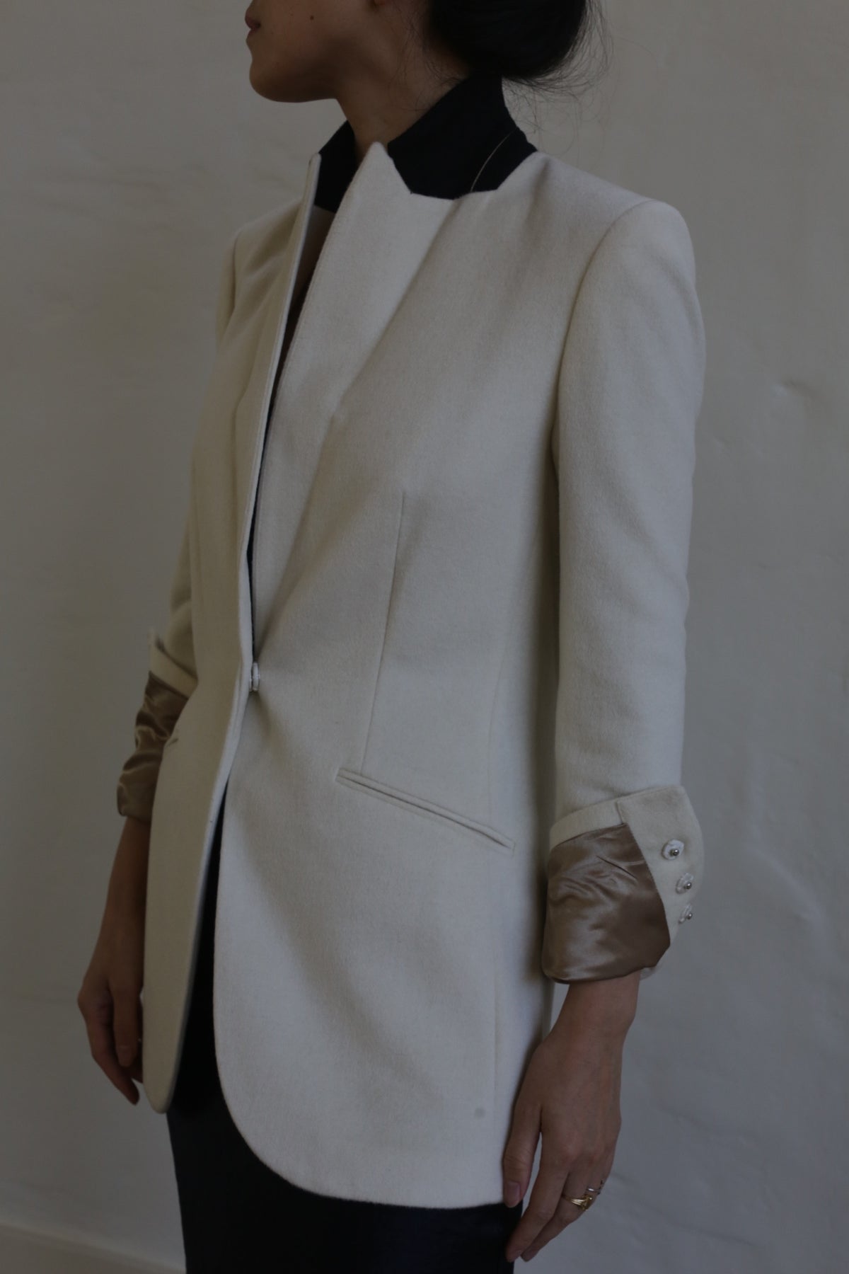 A person wearing the Sloane cashmere undyed blazer, tailored in a cream color with metallic sleeve cuffs, stands in profile against a plain backdrop. The blazer features a sleek design reminiscent of men's tailoring, complete with a concealed front closure and a side pocket.