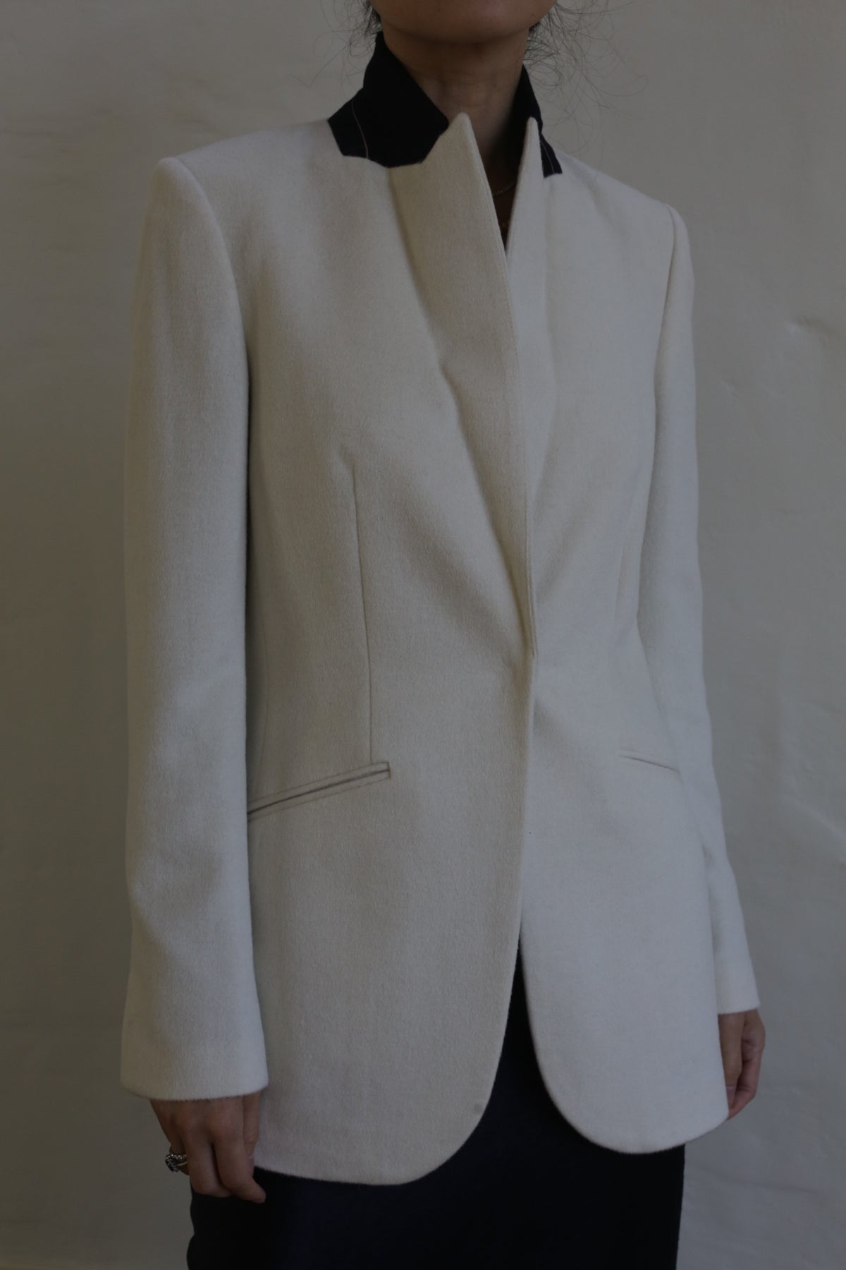 A person wearing the Sloane cashmere undyed blazer with a black collar stands against a neutral background. The tuxedo jacket features sleek, tailored lines and no visible buttons, adding a modern touch to the outfit. The person's face is not shown.