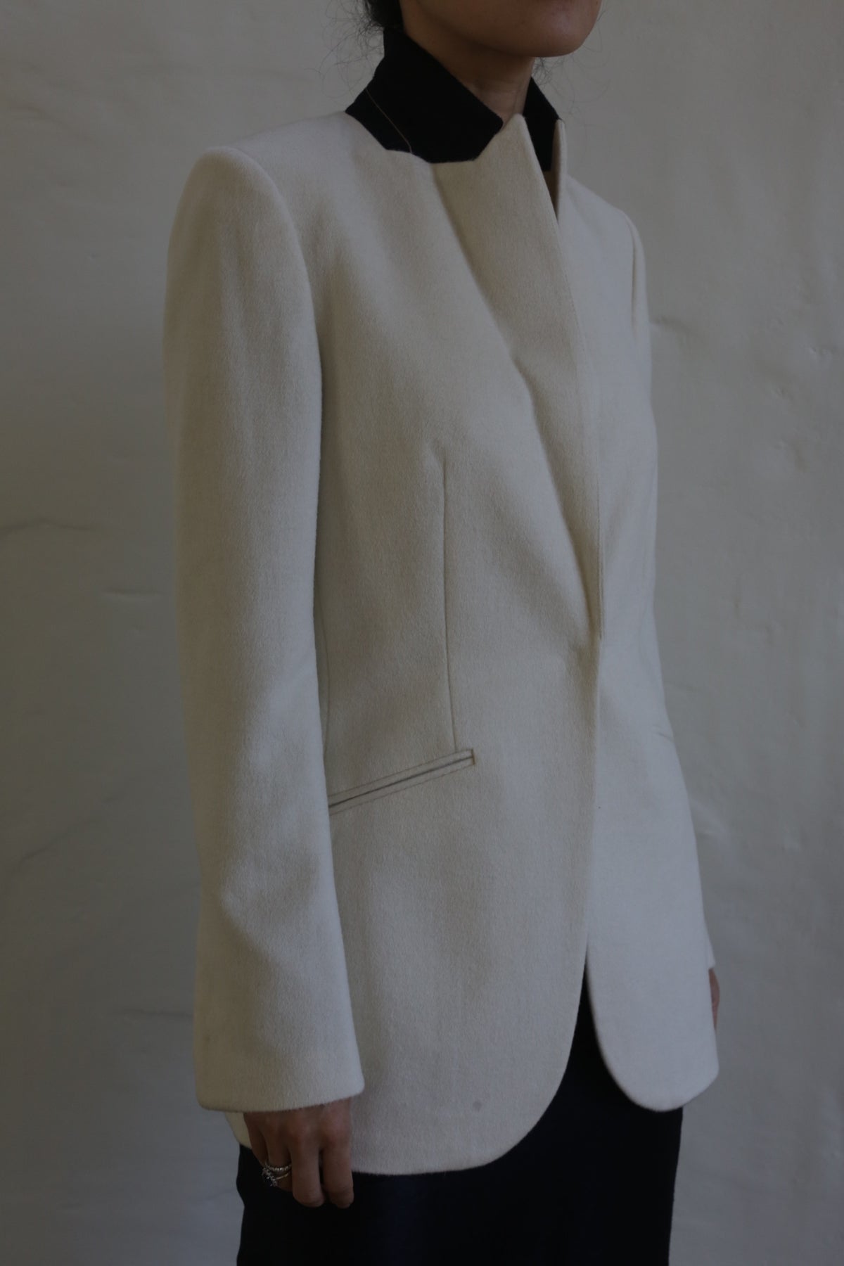 A person dressed in the Sloane cashmere undyed blazer stands against a neutral background, with their face partially out of view. The cream-colored jacket features a black collar reminiscent of a tuxedo, and it is paired with black skirt or pants. The blazer's design showcases simple, elegant lines that reflect the finesse of men's tailoring.