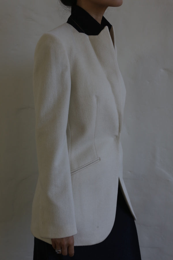 A person wearing the Sloane Cashmere Undyed blazer, which draws inspiration from classic men's tailoring, over a dark shirt is seen in profile against a neutral background. The image showcases the upper body and part of the skirt, while keeping the face out of view.