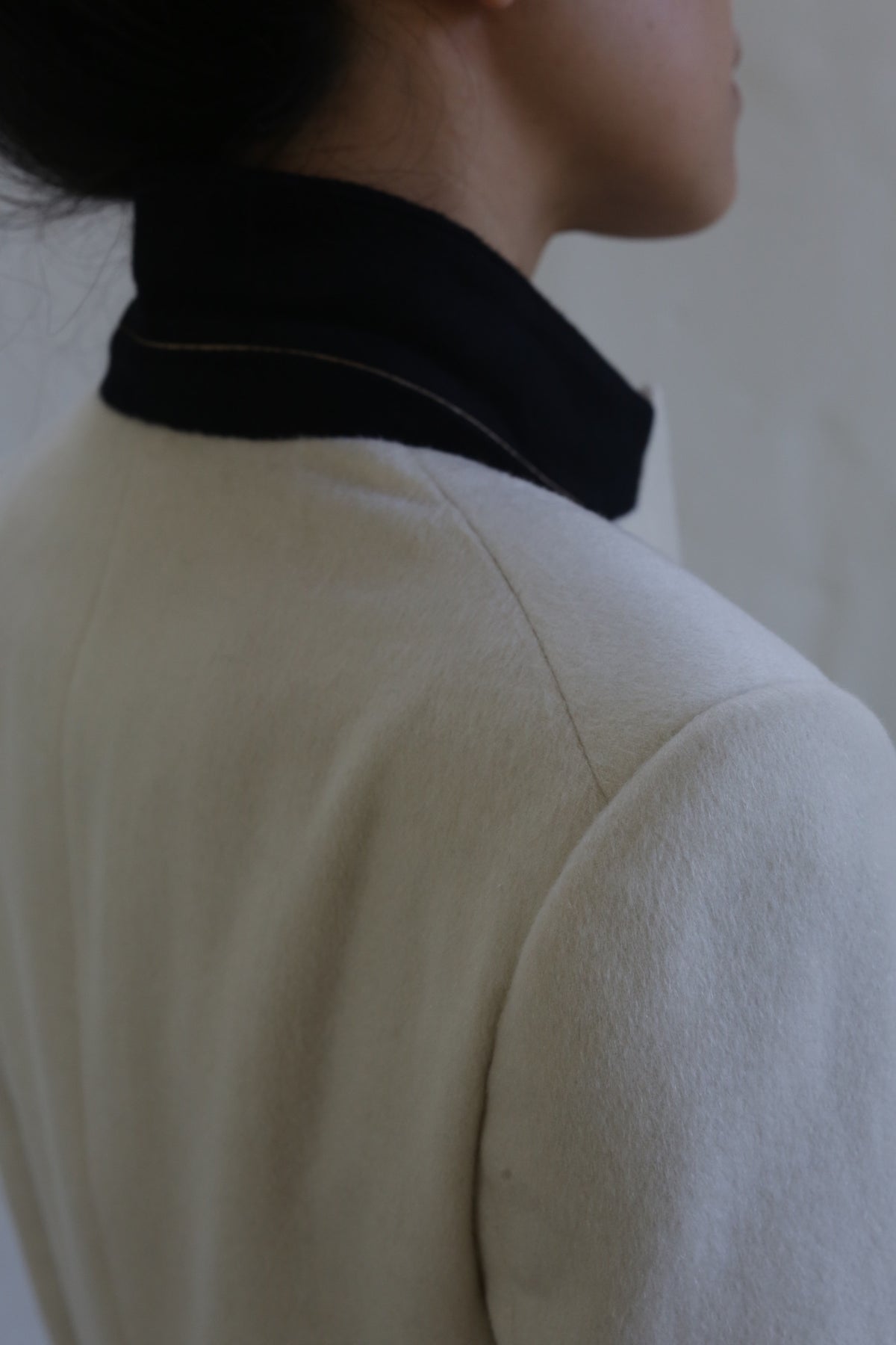 The image features a person viewed from behind, draped in the "Sloane | Cashmere Undyed" coat, which has a light hue paired with a contrasting dark collar. The focus on the shoulder and neck area enhances its minimalist and refined aesthetic, reminiscent of classic men's tailoring.