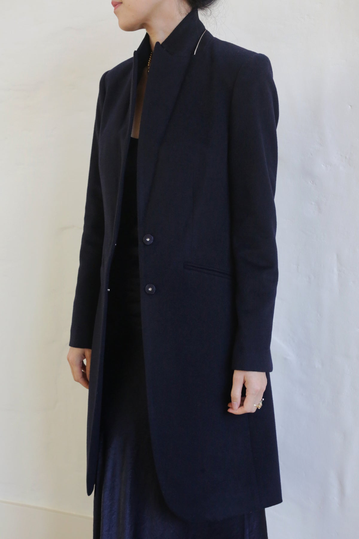 A person stands against a plain white background, wearing the Angeles | Navy, a long, dark-colored tuxedo coat made of 100% woven cashmere. The coat is buttoned at the waist and features a high collar. The person's face is turned away, and their hands rest at their sides.