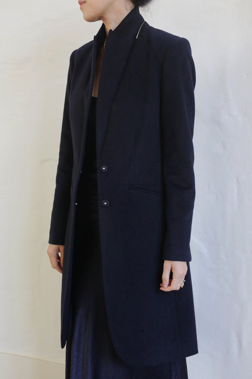 A person stands against a plain white background, wearing the Angeles | Navy, a long, dark-colored tuxedo coat made of 100% woven cashmere. The coat is buttoned at the waist and features a high collar. The person's face is turned away, and their hands rest at their sides.
