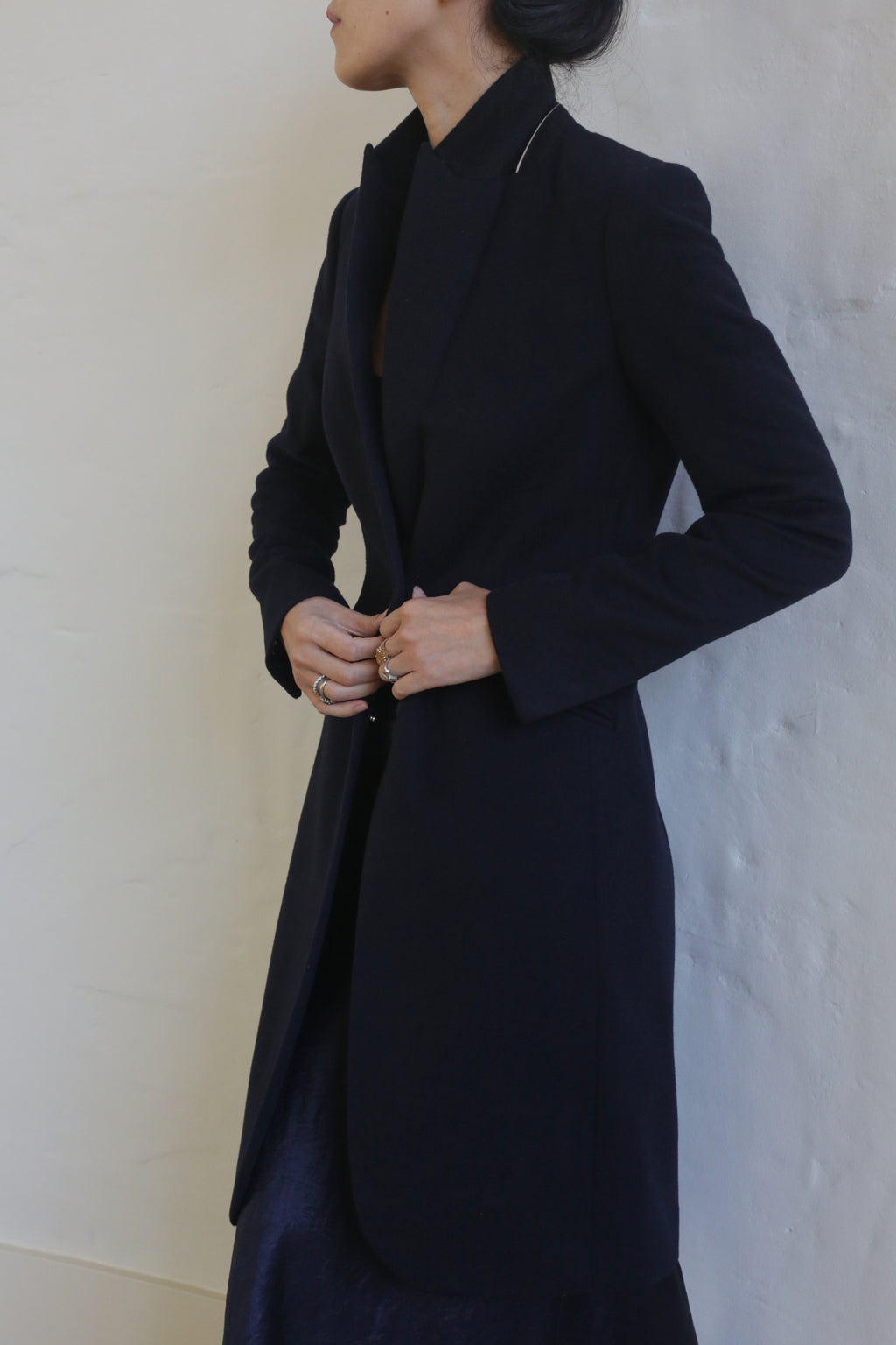 A person with long hair in an updo is wearing the Navy Angeles, a dark equestrian cut coat that falls to the knee, paired with a skirt. They are standing against a light-colored textured wall, buttoning the 100% woven cashmere coat with both hands. The person's face is turned to the side and is partially visible.