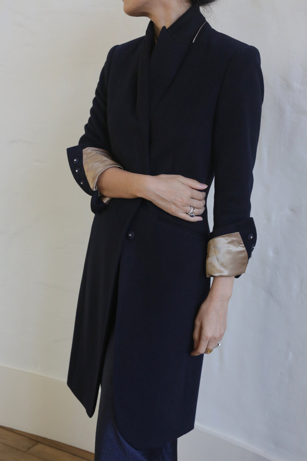 A person is shown from the neck down, wearing the Angeles | Navy, a dark-colored tailored equestrian cut blazer with light-colored cuffs folded up. The person has their arms crossed and is standing against a lightly textured, off-white wall. Rings are visible on the person's fingers.