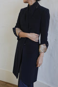 A person is shown from the neck down, wearing the Angeles | Navy, a dark-colored tailored equestrian cut blazer with light-colored cuffs folded up. The person has their arms crossed and is standing against a lightly textured, off-white wall. Rings are visible on the person's fingers.