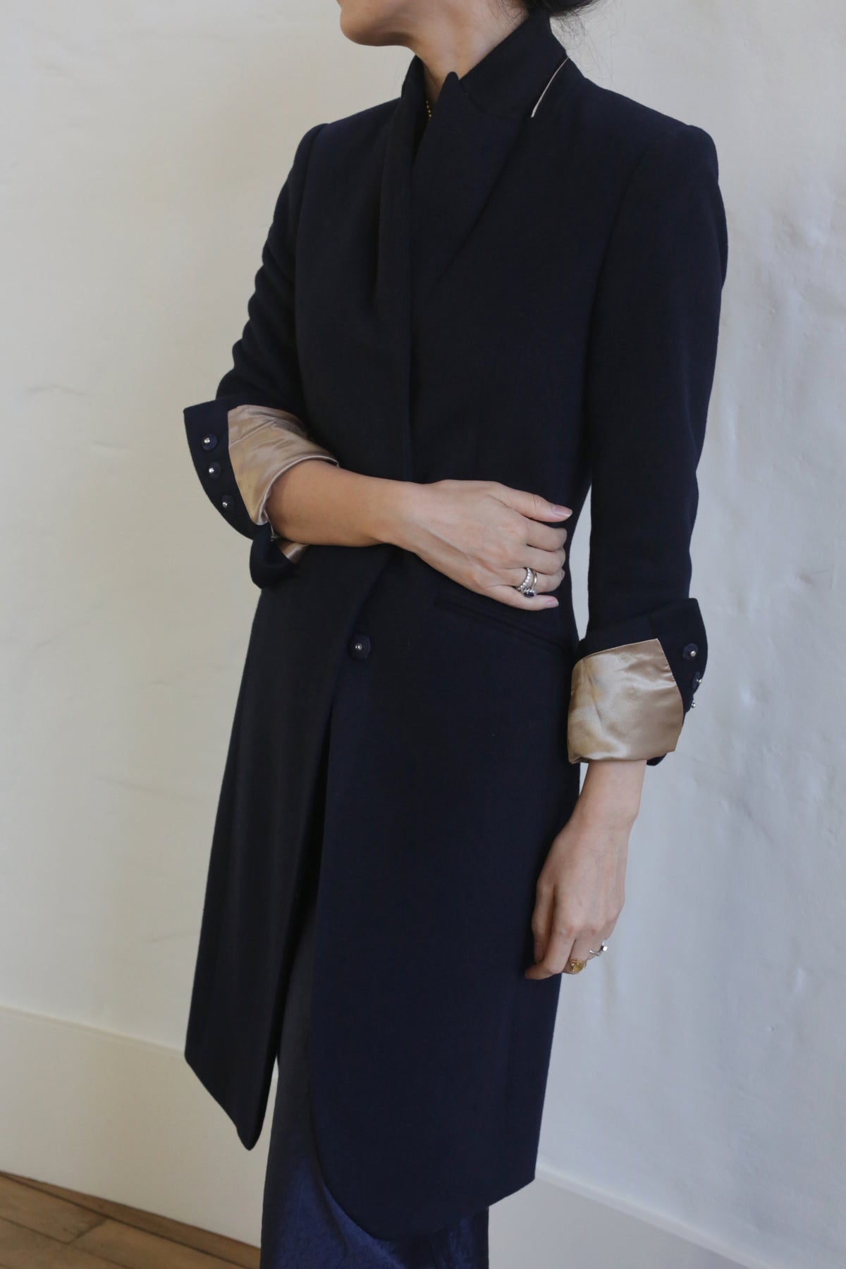 A person is shown from the neck down, wearing the Angeles | Navy, a dark-colored tailored equestrian cut blazer with light-colored cuffs folded up. The person has their arms crossed and is standing against a lightly textured, off-white wall. Rings are visible on the person's fingers.