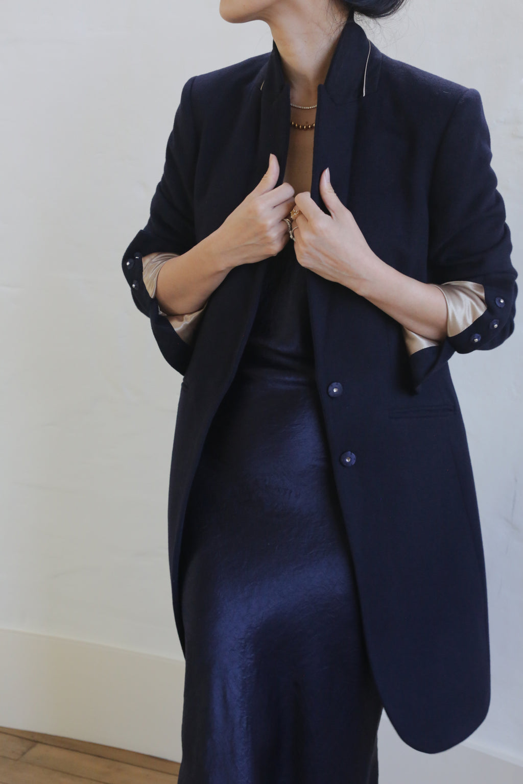 A person is shown from the neck down, wearing a dark, long Tuxedo coat with an equestrian cut. The sleeves are rolled up to reveal a shiny inner lining. They are also wearing a shiny, dark dress and holding the collar of the "Angeles | Navy" 100% woven cashmere coat with both hands. The background is a light-colored wall.