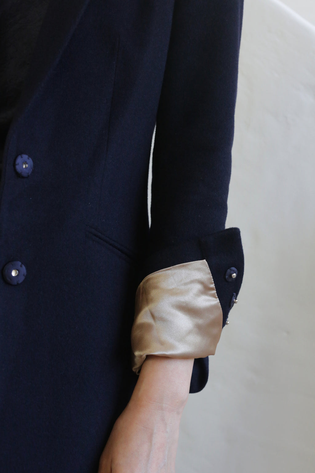 A person wearing the Angeles | Navy, a dark navy, 100% woven cashmere blazer with light gold satin lining visible as the sleeves are stylishly rolled up. The blazer features dark buttons on the sleeve and front. The background is neutral and out of focus.