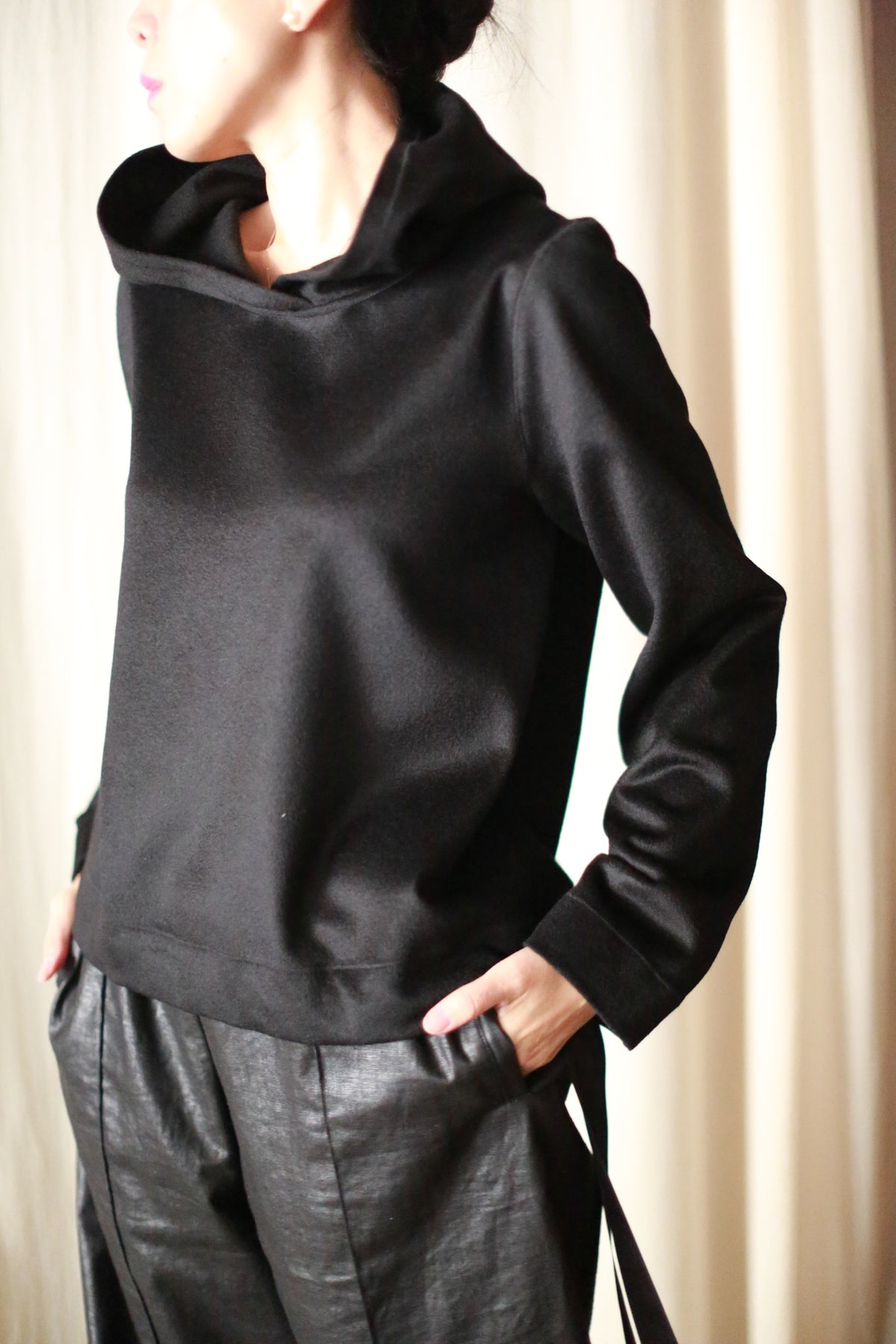 A person wears the Alex Hoodie in black, paired with black leather pants, standing with hands in their pockets. Soft lighting and beige curtains enhance the luxurious look of this carefully crafted ensemble.