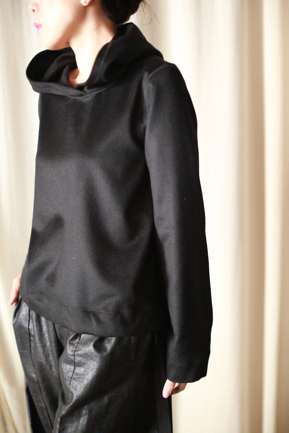 A person in an Alex Hoodie | Black and dark, textured pants stands against a light curtain, showcasing a modern minimalist style with soft shadows accentuating the luxurious attire.