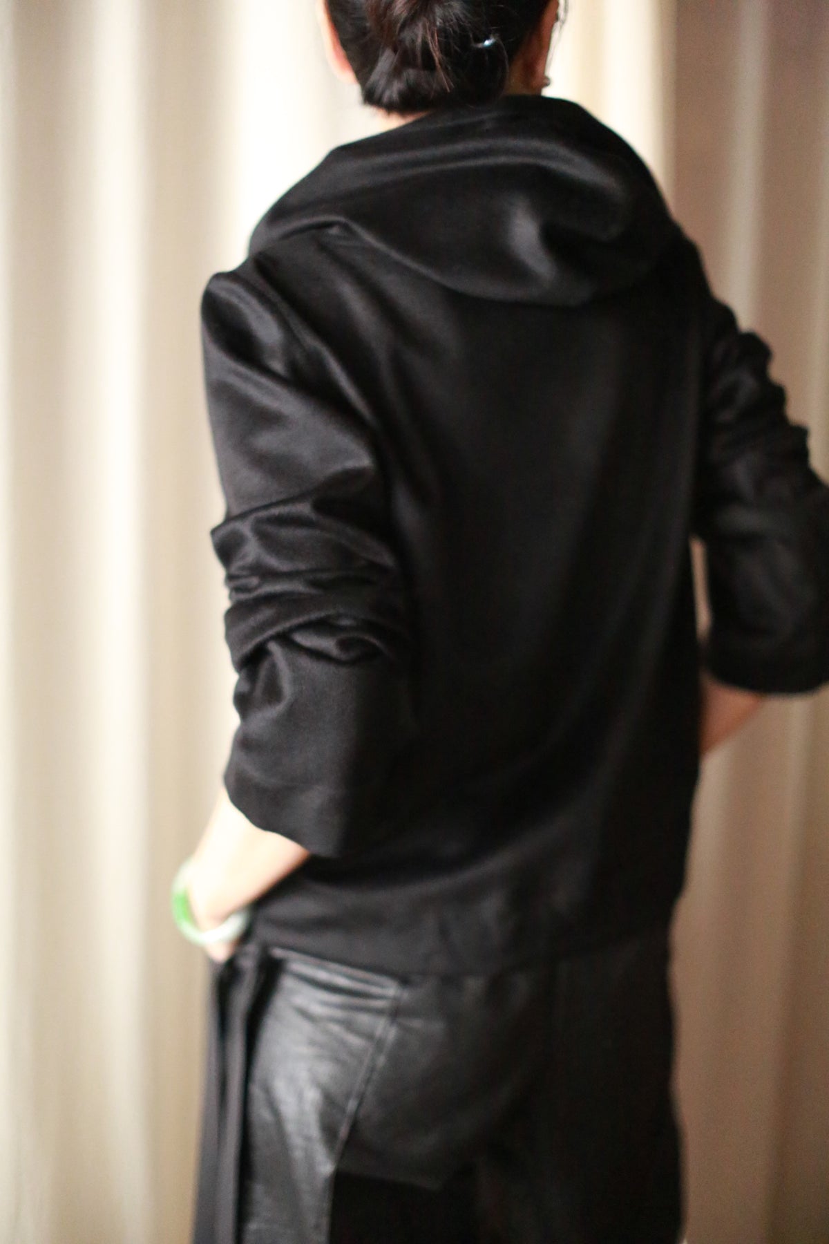 A dark-haired person in a bun, wearing the luxurious Alex Hoodie in black with leather pants, stands with hands on hips against a light-colored drape background.