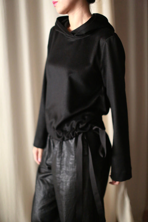 A person is elegantly dressed in a luxurious long-sleeve Alex Hoodie in black, paired with black pants, against a blurred cream curtain. Partially facing left, this handmade piece from San Francisco exudes understated elegance.