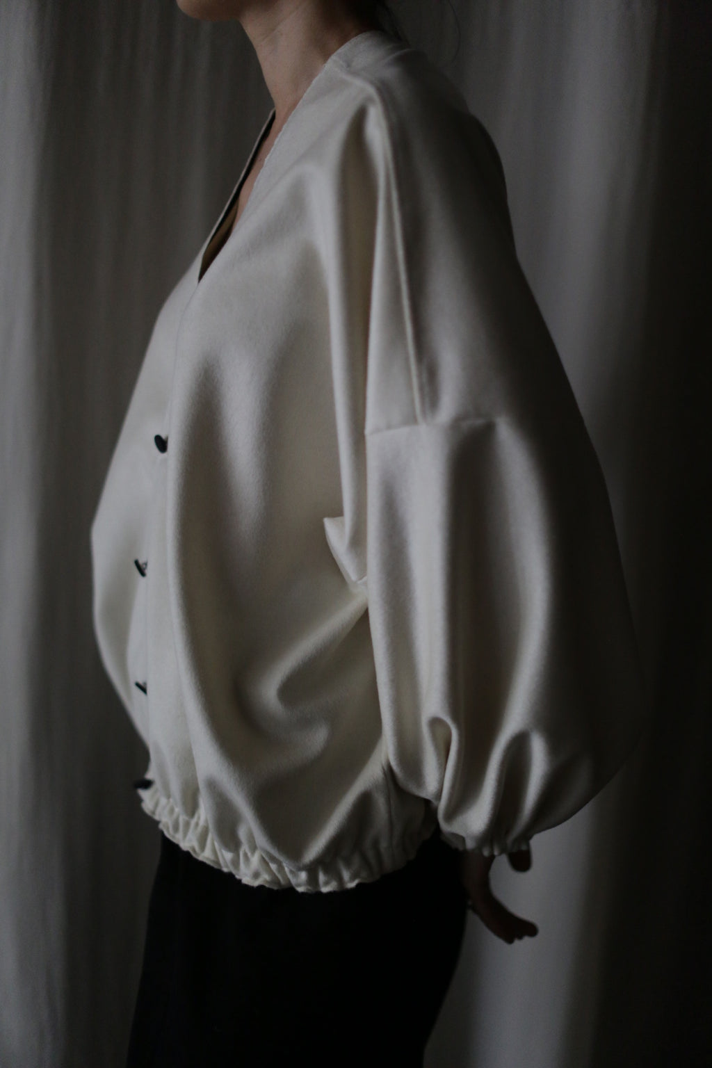 A person stands sideways against a dark background wearing "Blanc de Chine," a loose cream-colored long-sleeve blouse with black buttons and gathered hem, emphasizing zero-waste design principles in its thoughtful creation.