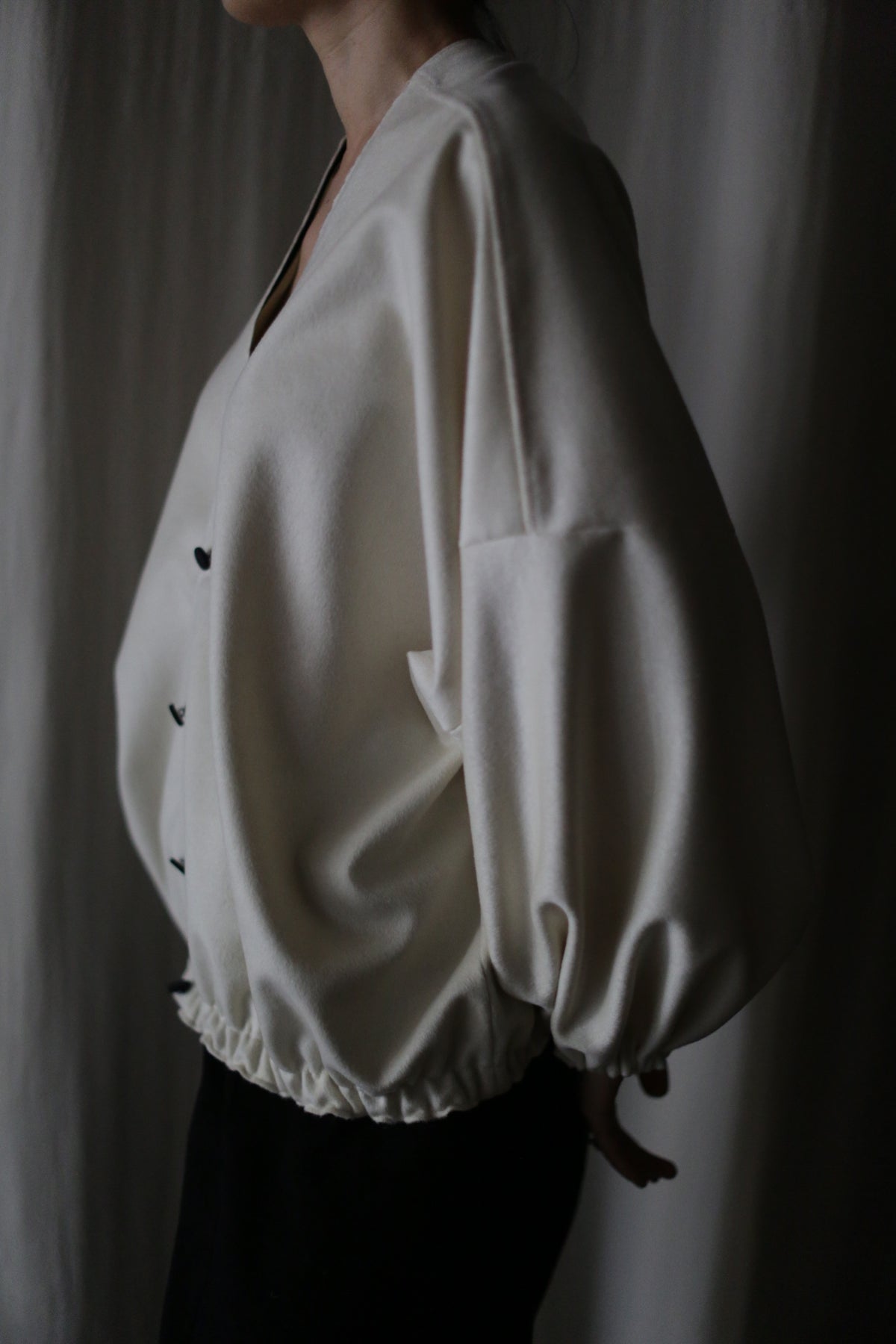 A person stands sideways against a dark background wearing "Blanc de Chine," a loose cream-colored long-sleeve blouse with black buttons and gathered hem, emphasizing zero-waste design principles in its thoughtful creation.