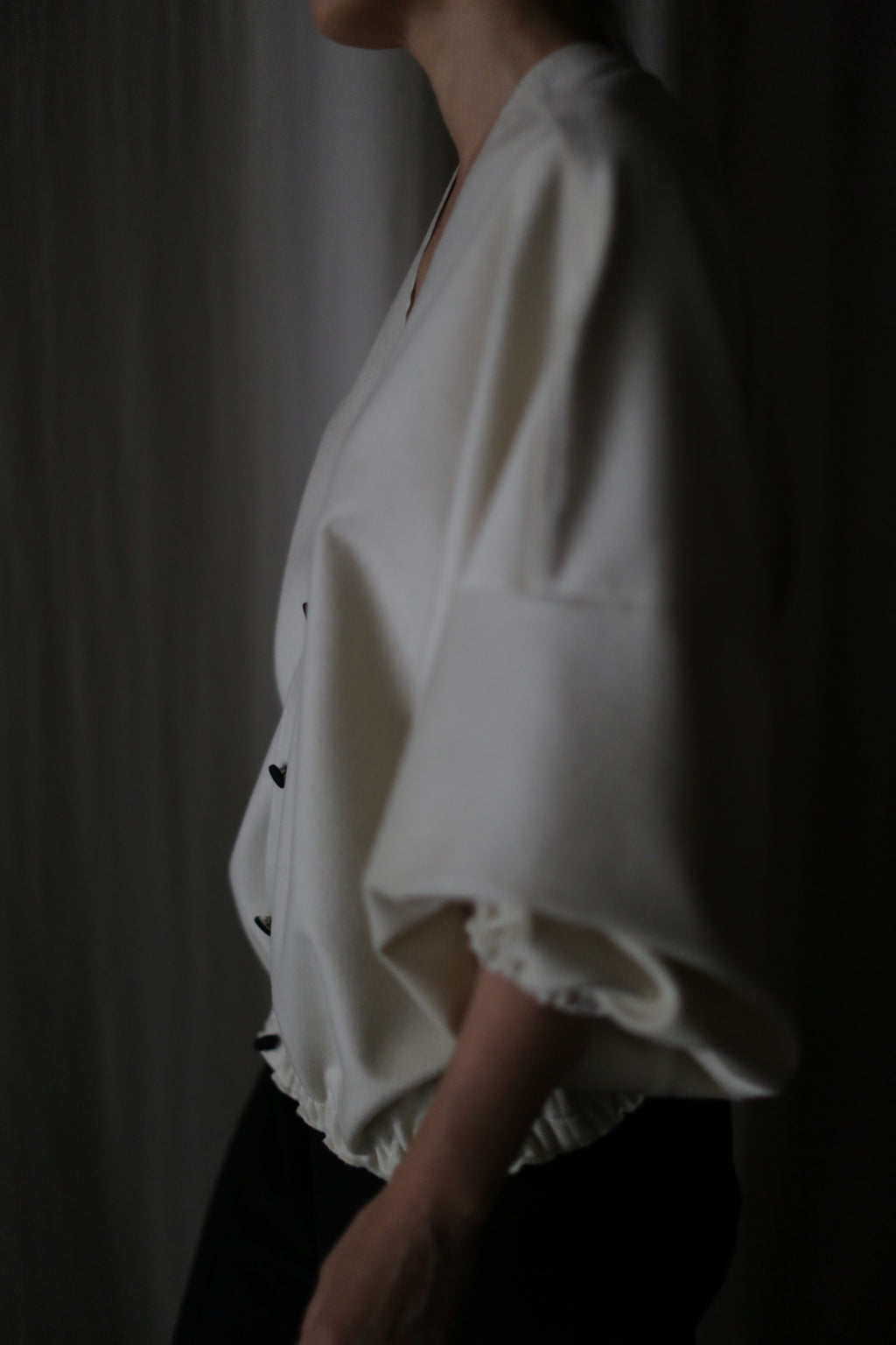 In dim lighting, a person showcases the Blanc de Chine: a loose-fitting white top with wide sleeves and black pants, highlighting its zero-waste design and commitment to sustainable fashion.