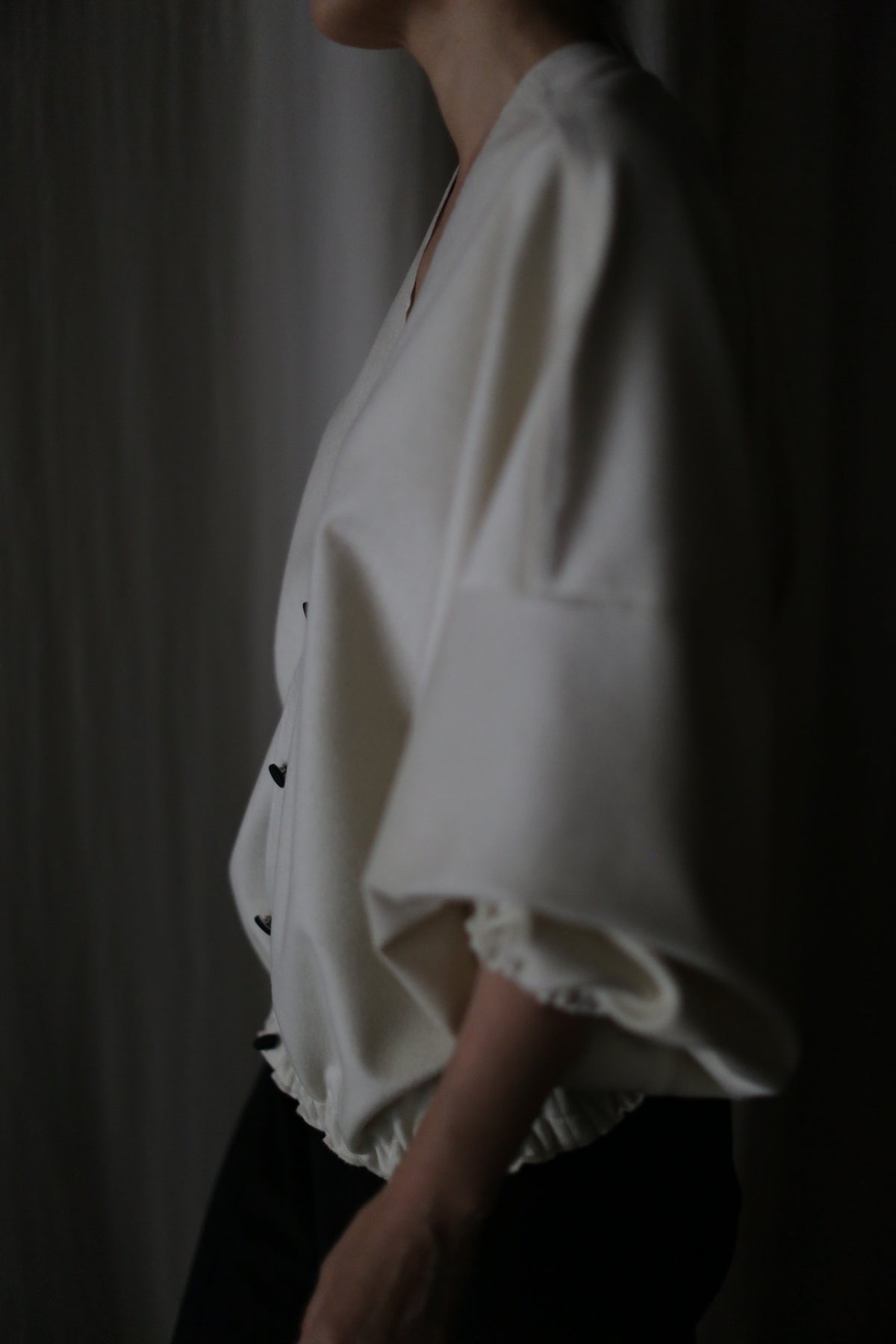 In dim lighting, a person showcases the Blanc de Chine: a loose-fitting white top with wide sleeves and black pants, highlighting its zero-waste design and commitment to sustainable fashion.