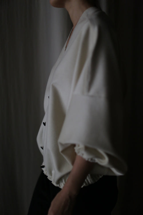 In dim lighting, a person showcases the Blanc de Chine: a loose-fitting white top with wide sleeves and black pants, highlighting its zero-waste design and commitment to sustainable fashion.