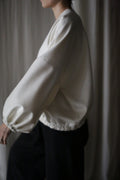 In a softly lit room, someone pairs dark pants with the Blanc de Chine blouse—featuring long sleeves and a loose fit designed on zero-waste principles. Their face is obscured by the neutral curtain backdrop, focusing attention on the sustainable fashion choice.