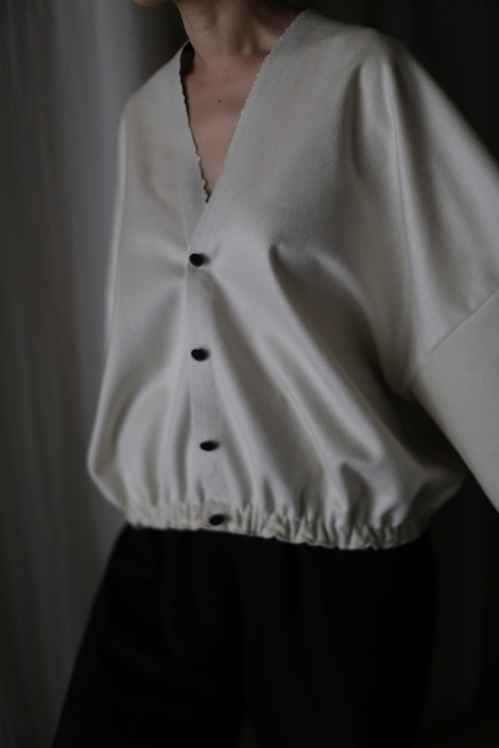 A person wears the Blanc de Chine, a handmade loose-fitting white cashmere top crafted in San Francisco. Featuring a deep V-neckline, long sleeves, black buttons, and an elastic waistband, it's styled with dark pants. The scene is softly lit against a dark background.