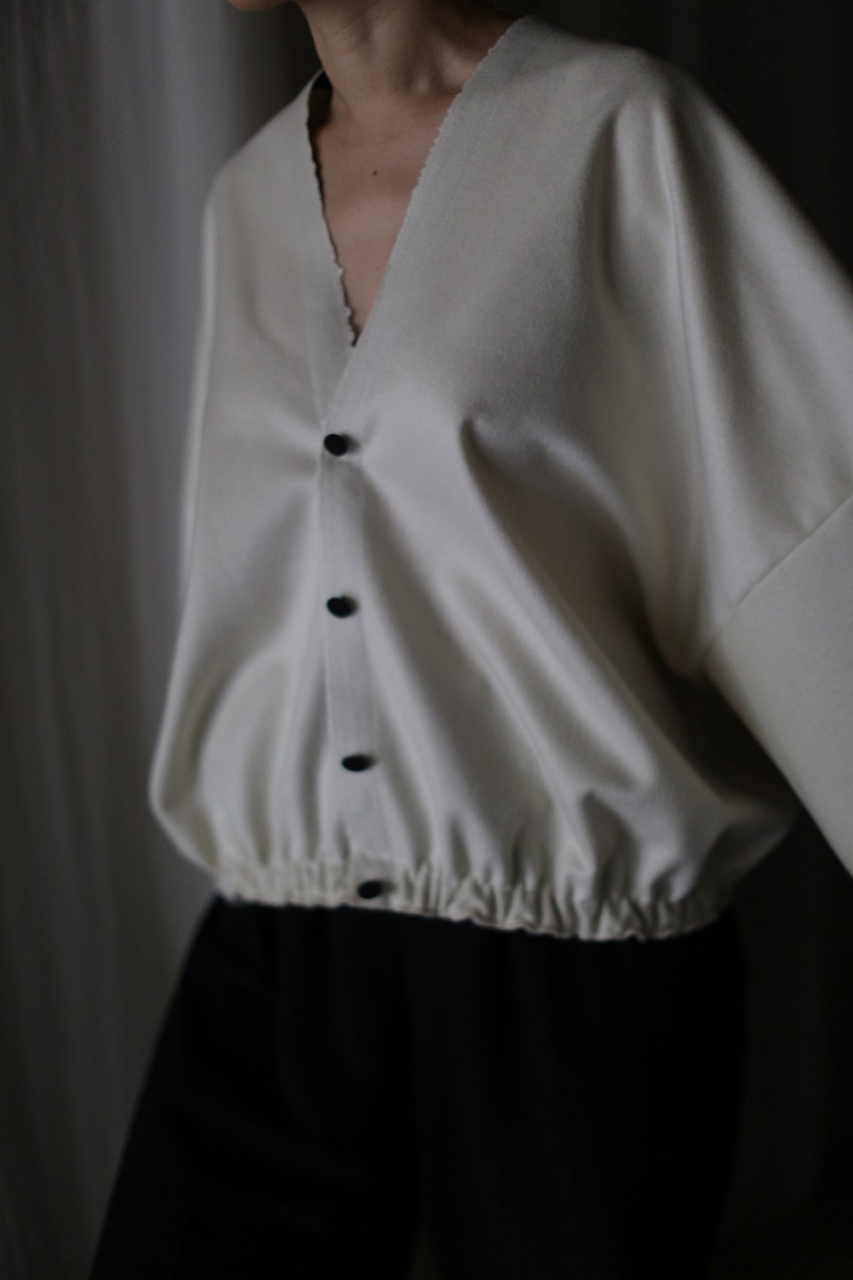 A person wears the Blanc de Chine, a handmade loose-fitting white cashmere top crafted in San Francisco. Featuring a deep V-neckline, long sleeves, black buttons, and an elastic waistband, it's styled with dark pants. The scene is softly lit against a dark background.