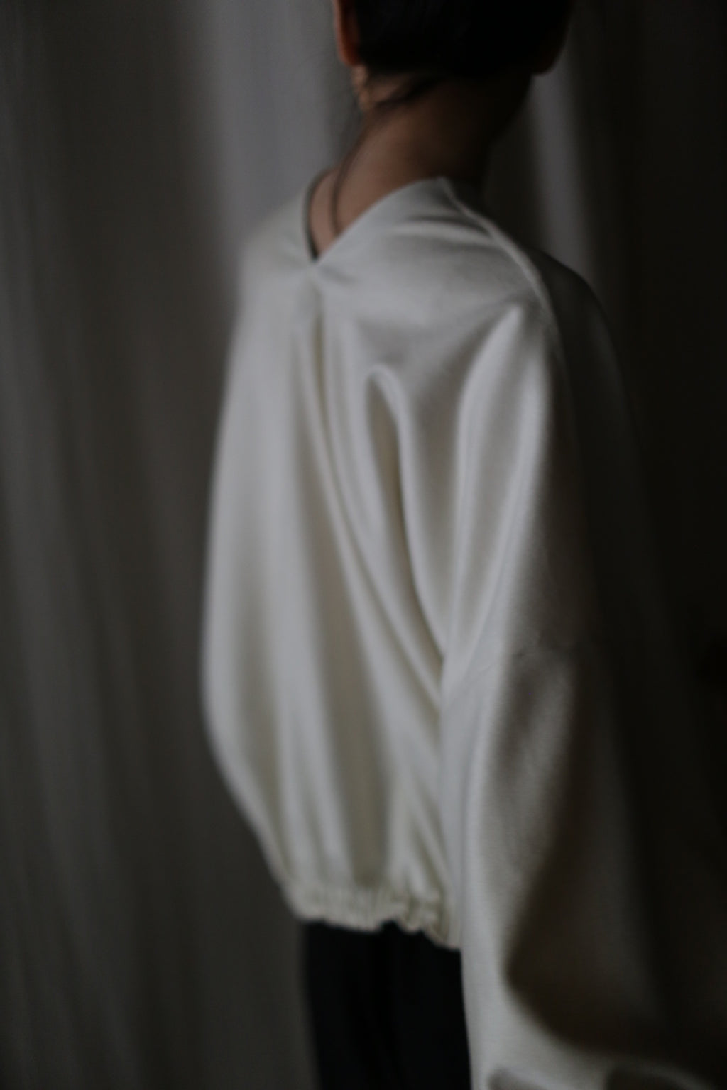 A person wearing a loose, light-colored Blanc de Chine blouse stands with their back to the camera against a softly lit background. The image is slightly out of focus, creating a serene atmosphere that highlights the elegance of zero-waste design.