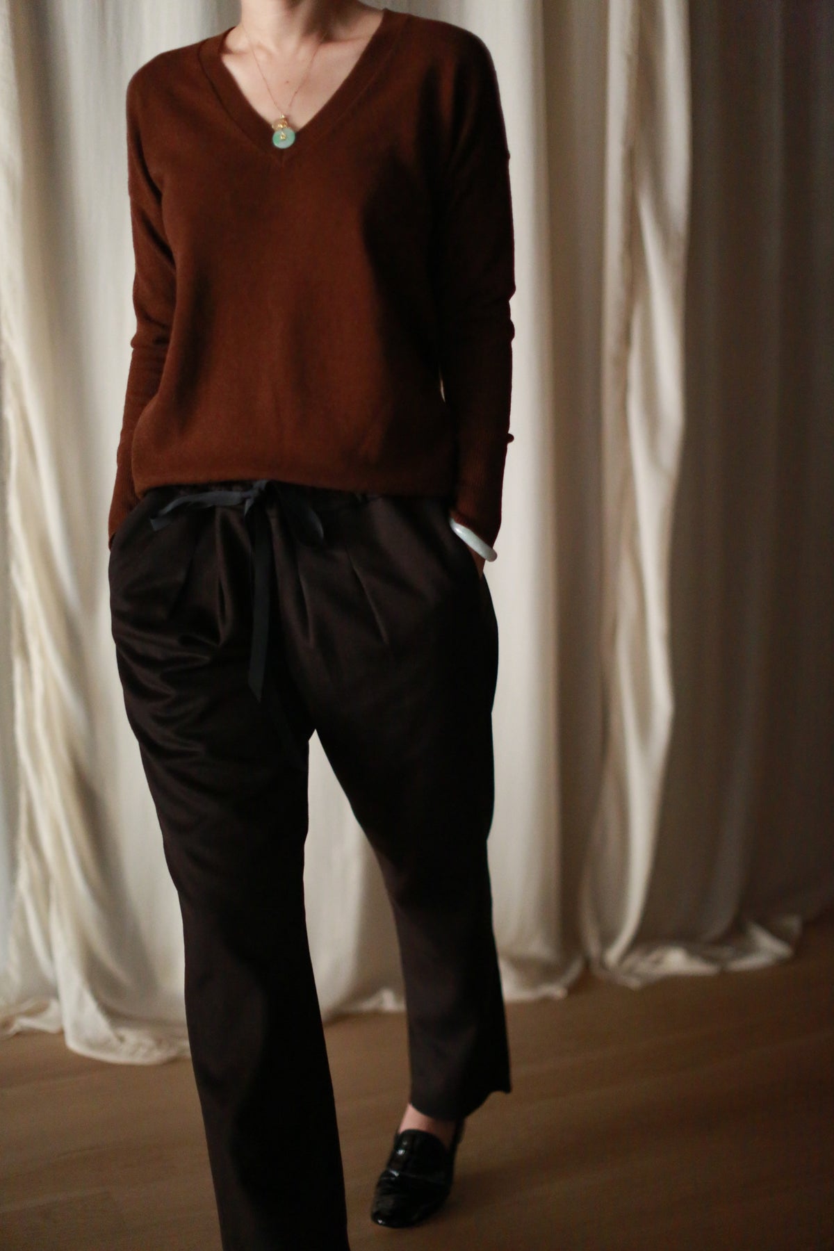 A person in a brown V-neck sweater and Cashmere Unisex Pants | Brown with a tie-waist detail stands with hands in their pockets. They are also wearing black shoes. The background features light draped curtains and a wooden floor. The person's face is not visible.