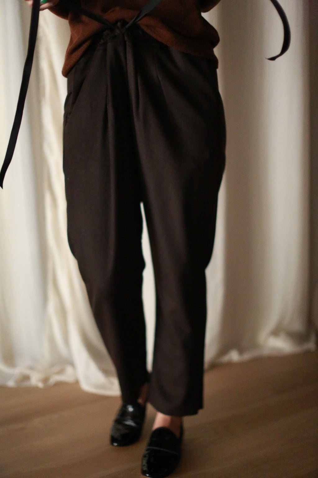 Person wearing Cashmere Unisex Pants | Brown tied with a silk ribbon drawstring, paired with black shoes. The background features light-colored, slightly wrinkled curtains. Only the lower half of the body is visible.