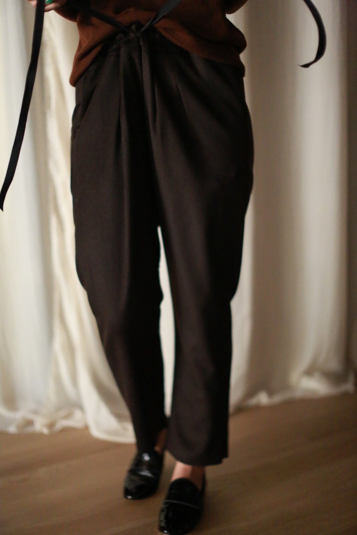 Person wearing Cashmere Unisex Pants | Brown tied with a silk ribbon drawstring, paired with black shoes. The background features light-colored, slightly wrinkled curtains. Only the lower half of the body is visible.