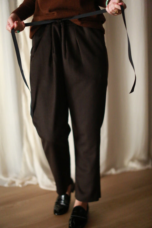 A person is shown tying a silk ribbon drawstring on Cashmere Unisex Pants | Brown. The background features soft white curtains, and the person is wearing a brown top and black shoes. Only the lower half of the body is visible, highlighting the unisex appeal of the outfit.