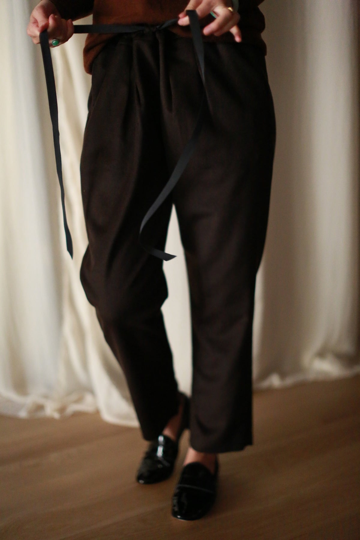 A person wearing Cashmere Unisex Pants | Brown with a silk ribbon drawstring and black shoes stands on a wooden floor in front of light-colored curtains. The individual is visible from the waist down, and their hands are holding the ribbon.