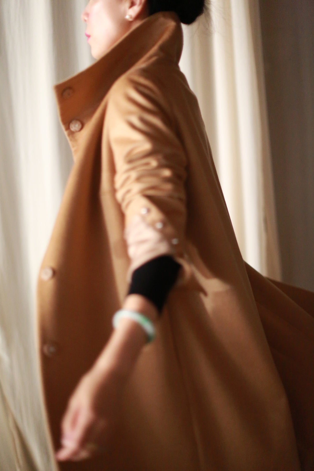 A person wearing the luxurious Stamford Long, a cashmere coat with a high collar, is captured mid-motion. The coat's button details on the cuffs add elegance. Softly lit cream-colored drapery forms the background, highlighting the fabric's texture and graceful movement.