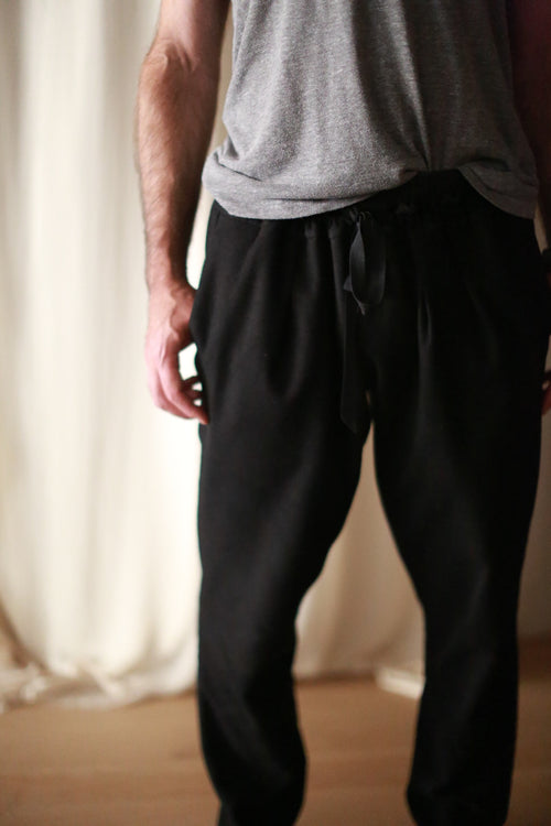 A person wearing a gray t-shirt and Cashmere Unisex Pants | Black is standing with their arms relaxed at their sides. The background is softly lit with light-colored curtains and a wooden floor. The person's face is not visible in the image.