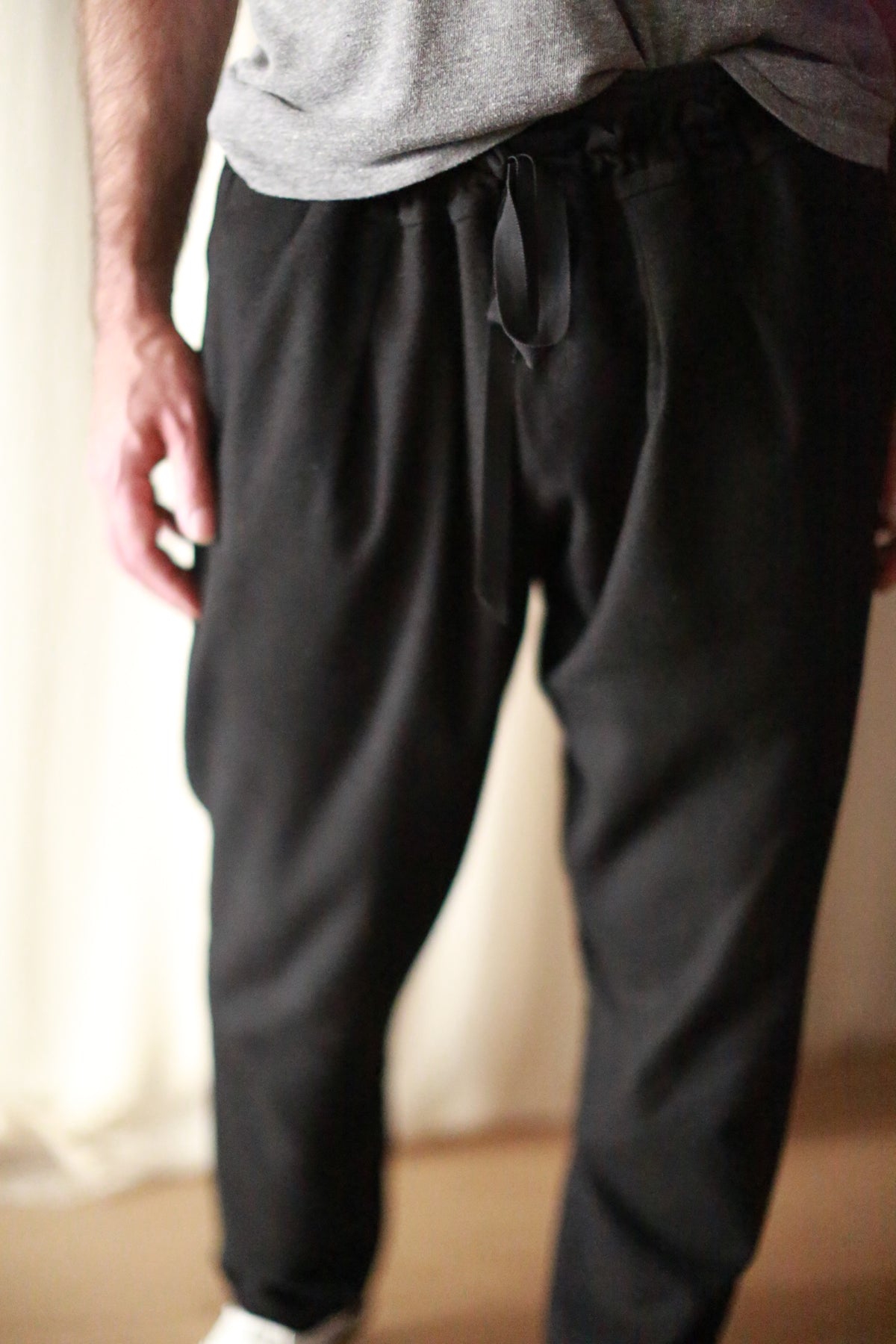A person wearing Cashmere Unisex Pants | Black with a silk ribbon drawstring and a gray shirt is shown from the neck down. The hands are relaxed at the sides, and the background appears to be neutral-colored curtains. The image focuses on the lower half of the body, highlighting the unisex design.