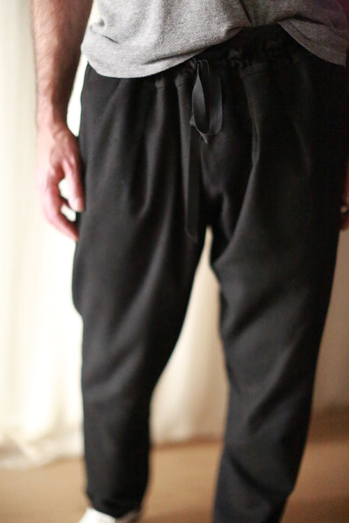 A person wearing Cashmere Unisex Pants | Black with a silk ribbon drawstring and a gray shirt is shown from the neck down. The hands are relaxed at the sides, and the background appears to be neutral-colored curtains. The image focuses on the lower half of the body, highlighting the unisex design.